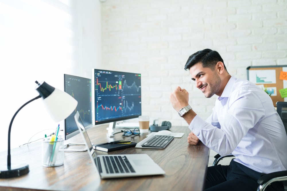 handsome swing trader sitting in front of his computer monitors happy and with his fist raised